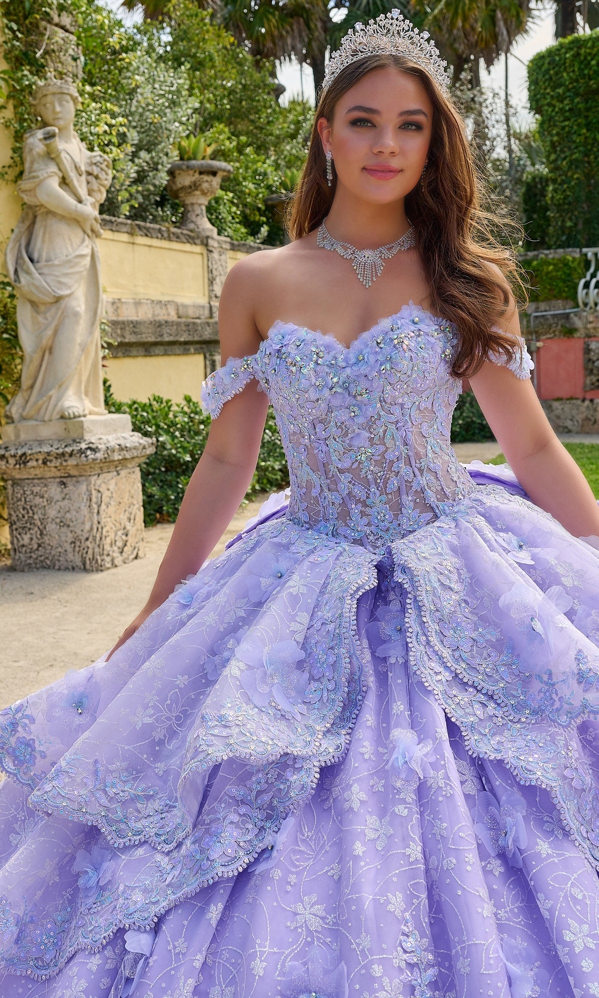 A young woman wears the Quinceanera Dress 54202 by Amarra—an off-the-shoulder ball gown with lace embroidered flowers and a tiara—while posing outdoors by a stone statue and garden, looking at the camera.